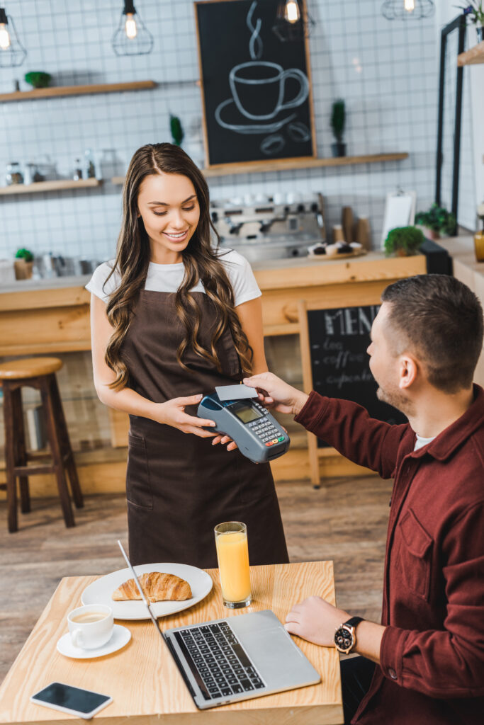 attractive waitress holding terminal wile handsome man sitting at table with laptop and paying with