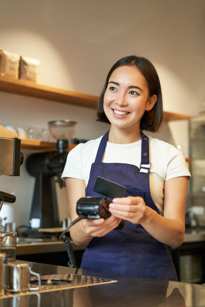 Portrait of smiling asian barista, coffee shop employee using POS terminal and credit card, helps